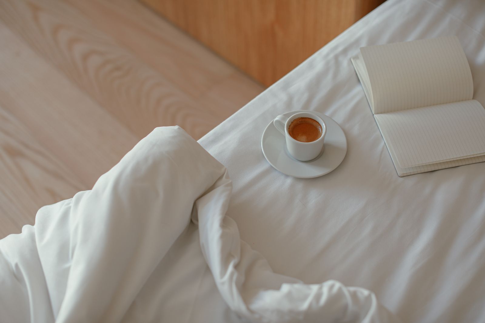 Un carnet ouvert et une tasse de café posés sur un lit aux draps blancs, dans une chambre au parquet clair