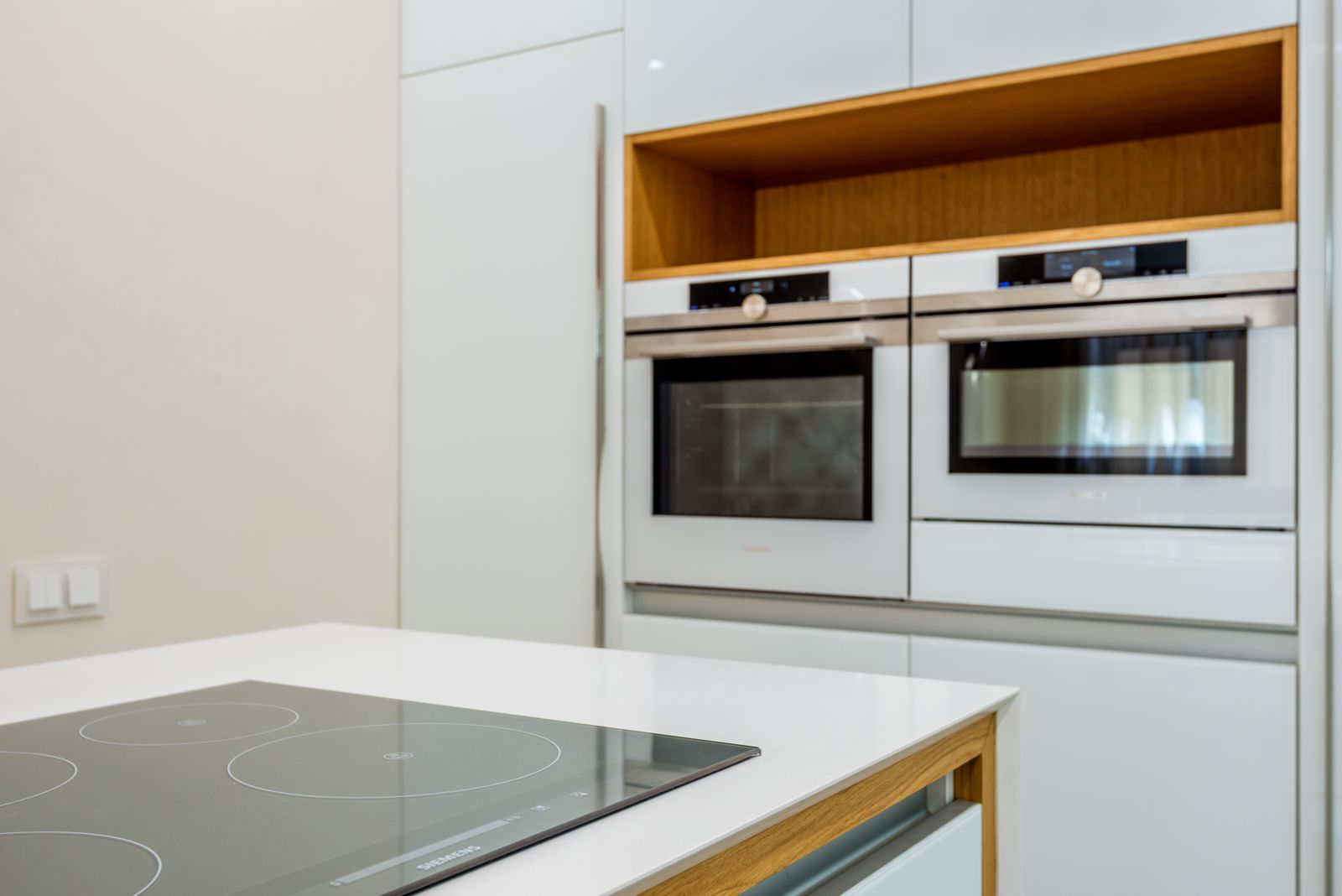 Une cuisine blanche et lumineuse avec une plaque à induction au premier plan et deux fours encastrés dans une niche en bois