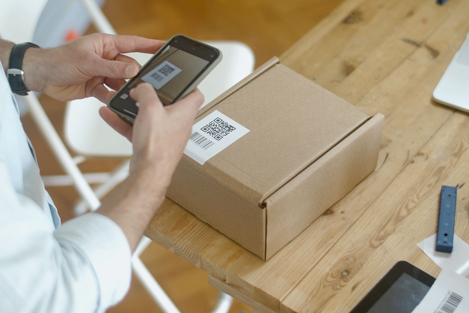 Deux mains tiennent un téléphone, appareil photo ouvert, au-dessus d'un carré noir et blanc collé sur un colis posé sur une table en bois clair