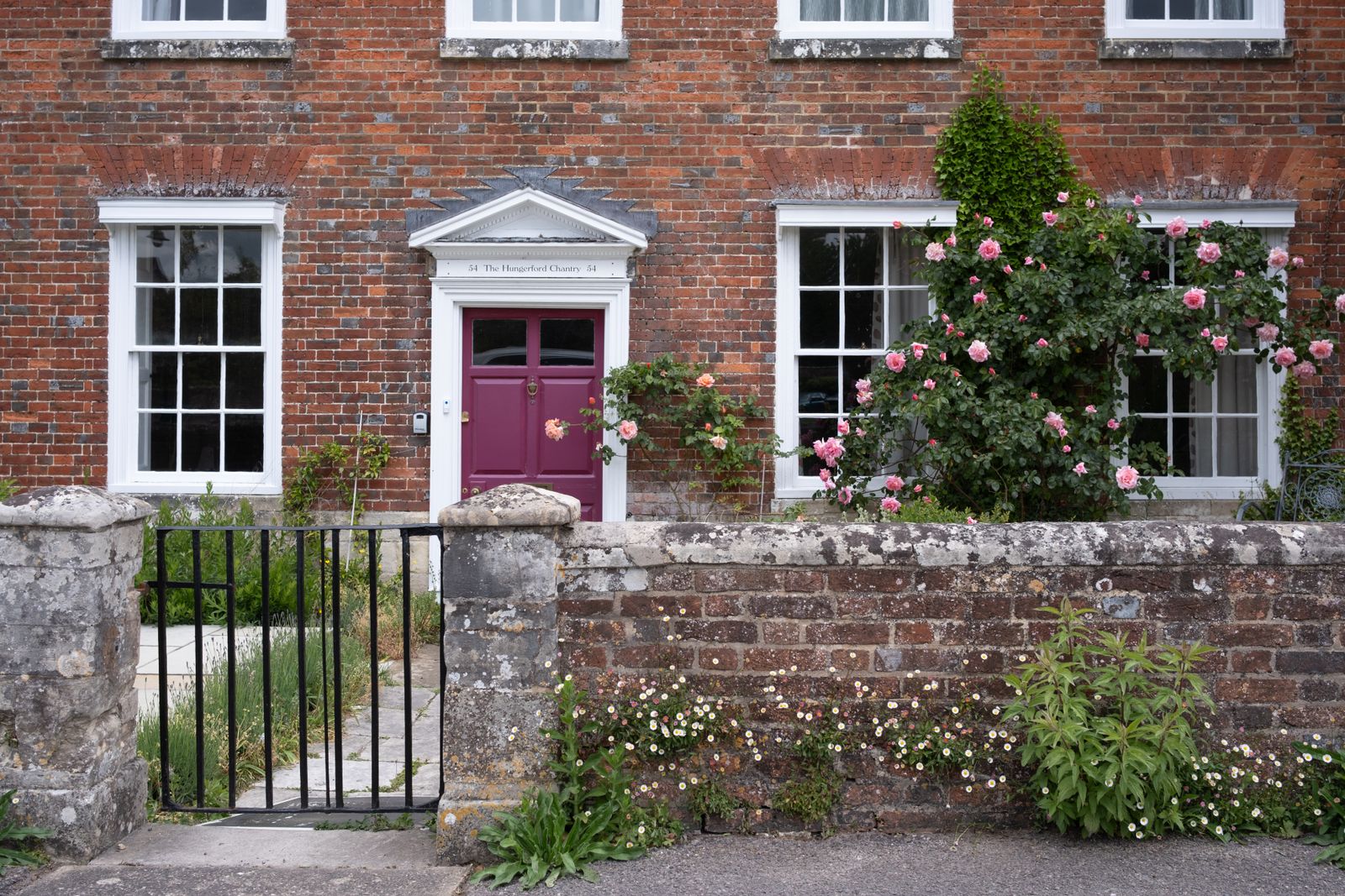 Un portail en fer ouvert sur une allée dallée qui mène à la porte prune d'une maison de briques, des rosiers en fleur sous les fenêtres