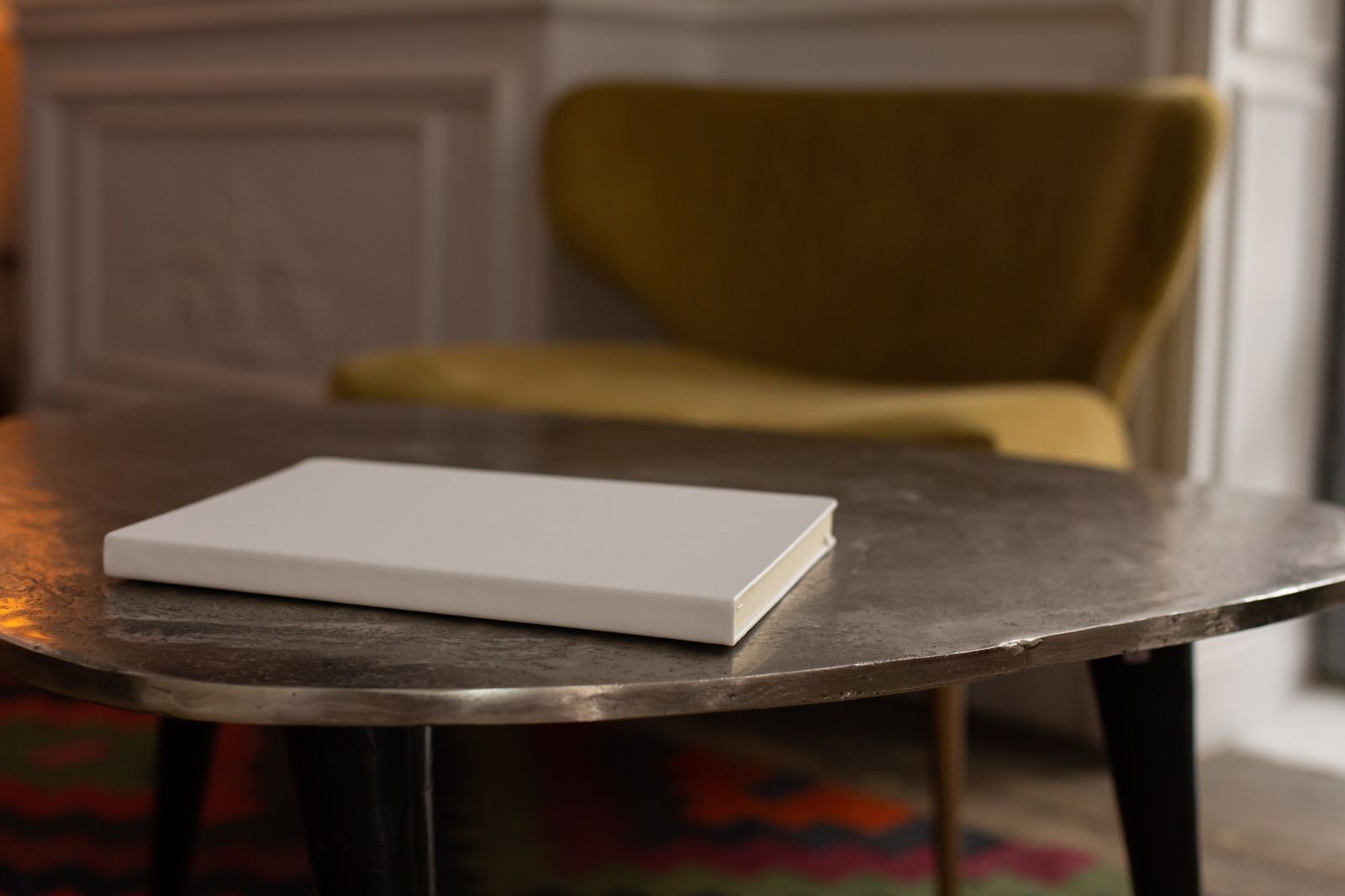 Un livret blanc fermé posé sur une table basse, devant un fauteuil en velours moutarde dans un salon