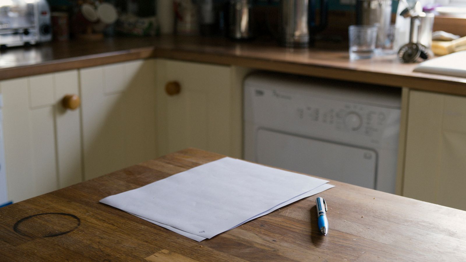 Une feuille blanche et un stylo posés sur la table en bois d'une cuisine aux placards crème, un lave-linge en arrière-plan
