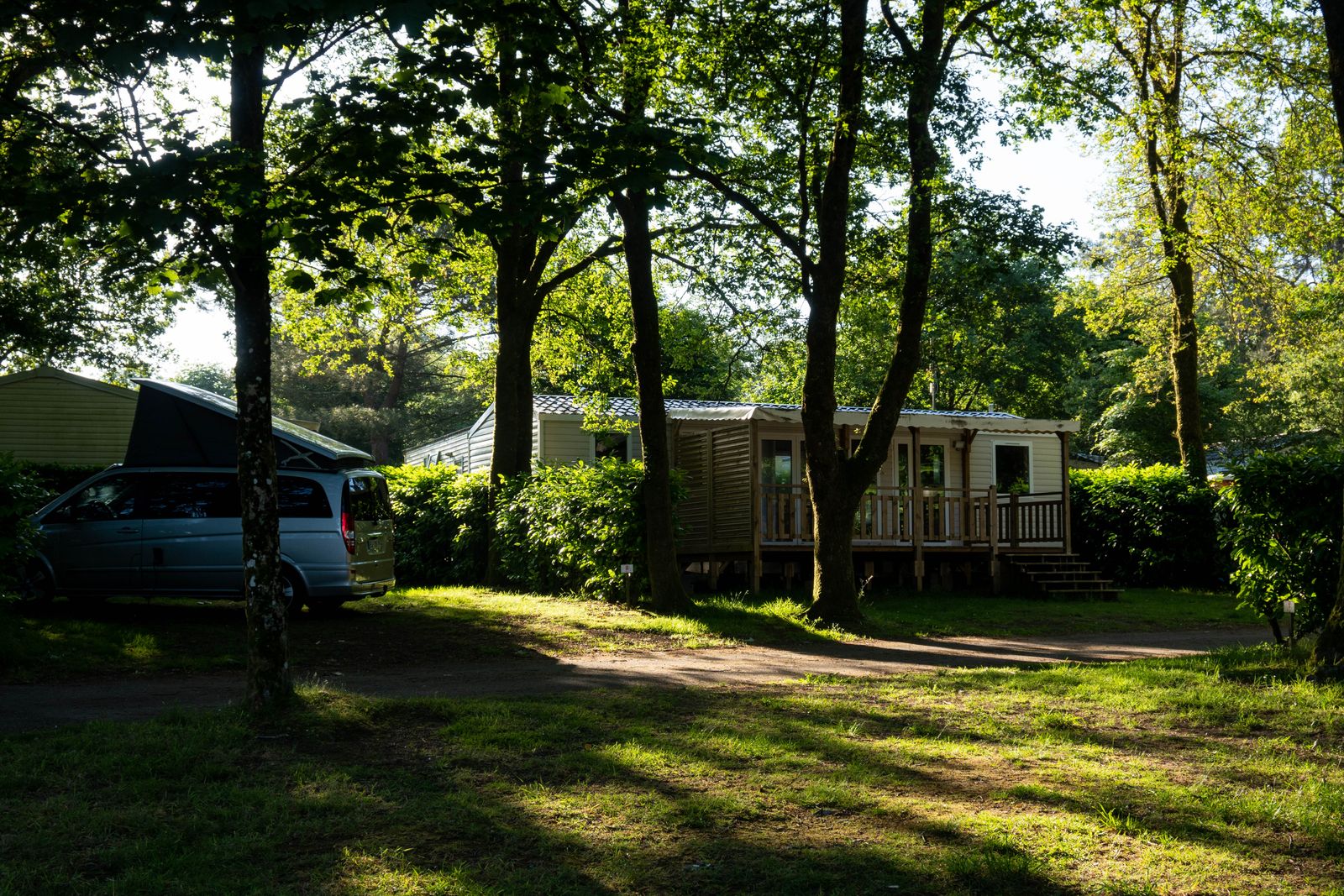 Un mobil-home avec sa terrasse en bois sous les arbres d'un camping, un van garé sur l'emplacement voisin en fin de journée