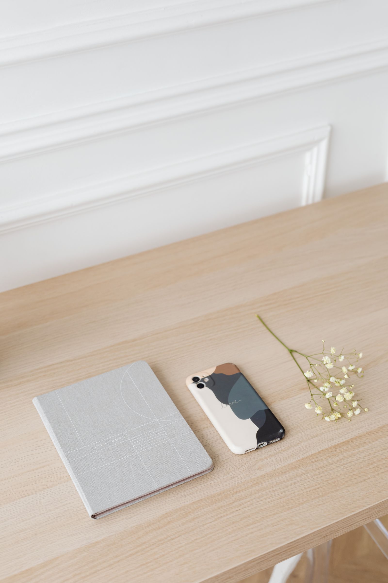 Un téléphone à coque colorée, un carnet gris et des fleurs blanches sur une table en bois clair