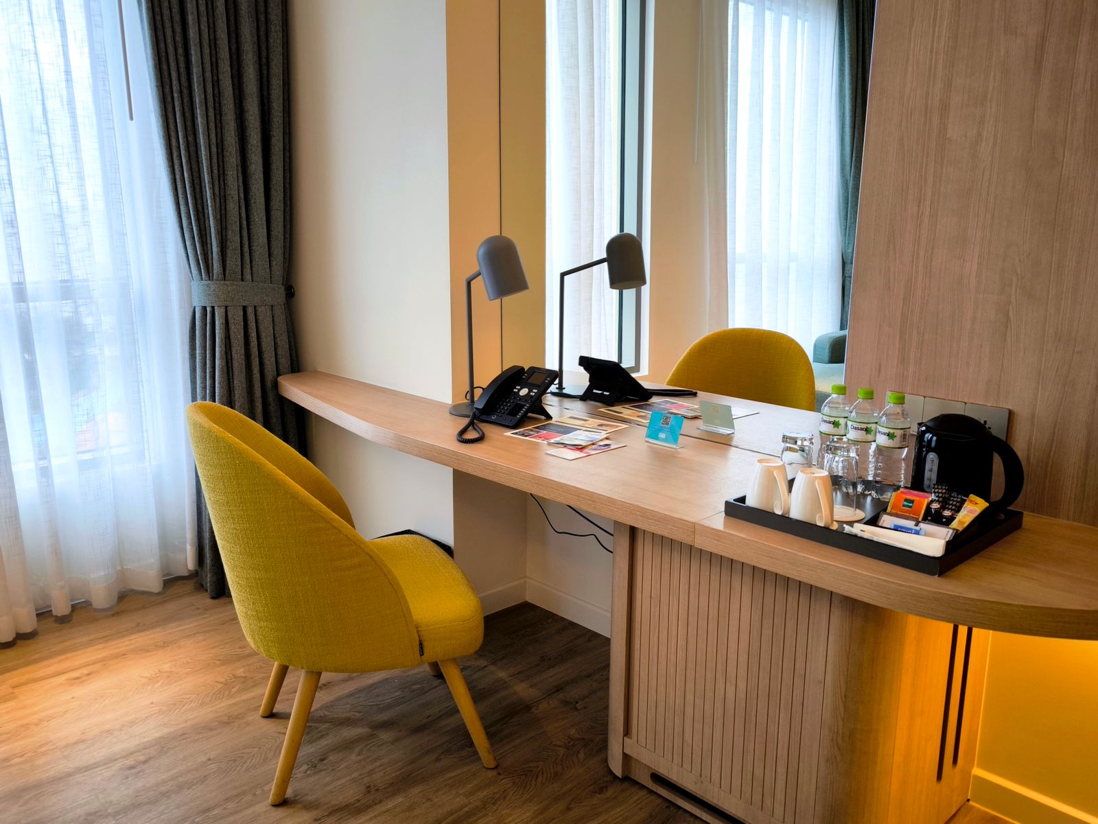 Le bureau en bois clair d'une chambre d'hôtel, avec téléphone, lampe, plateau de courtoisie et fauteuil jaune devant une grande fenêtre voilée