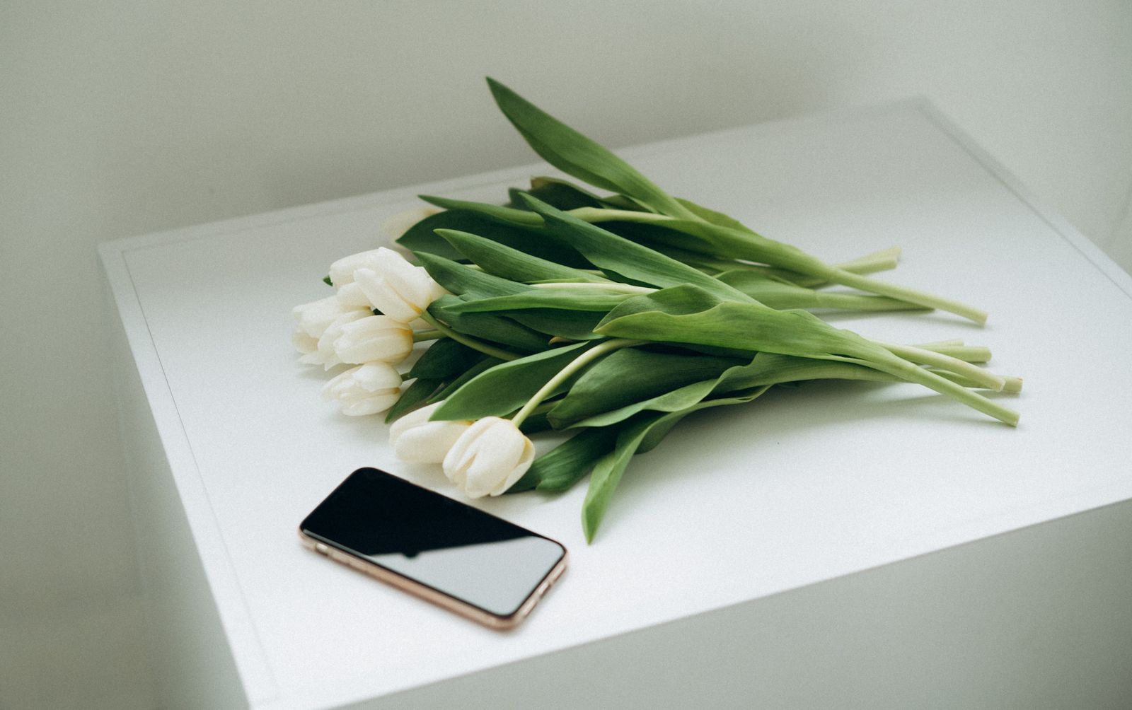 Un bouquet de tulipes blanches posé sur une table blanche, un téléphone à l'écran éteint juste à côté