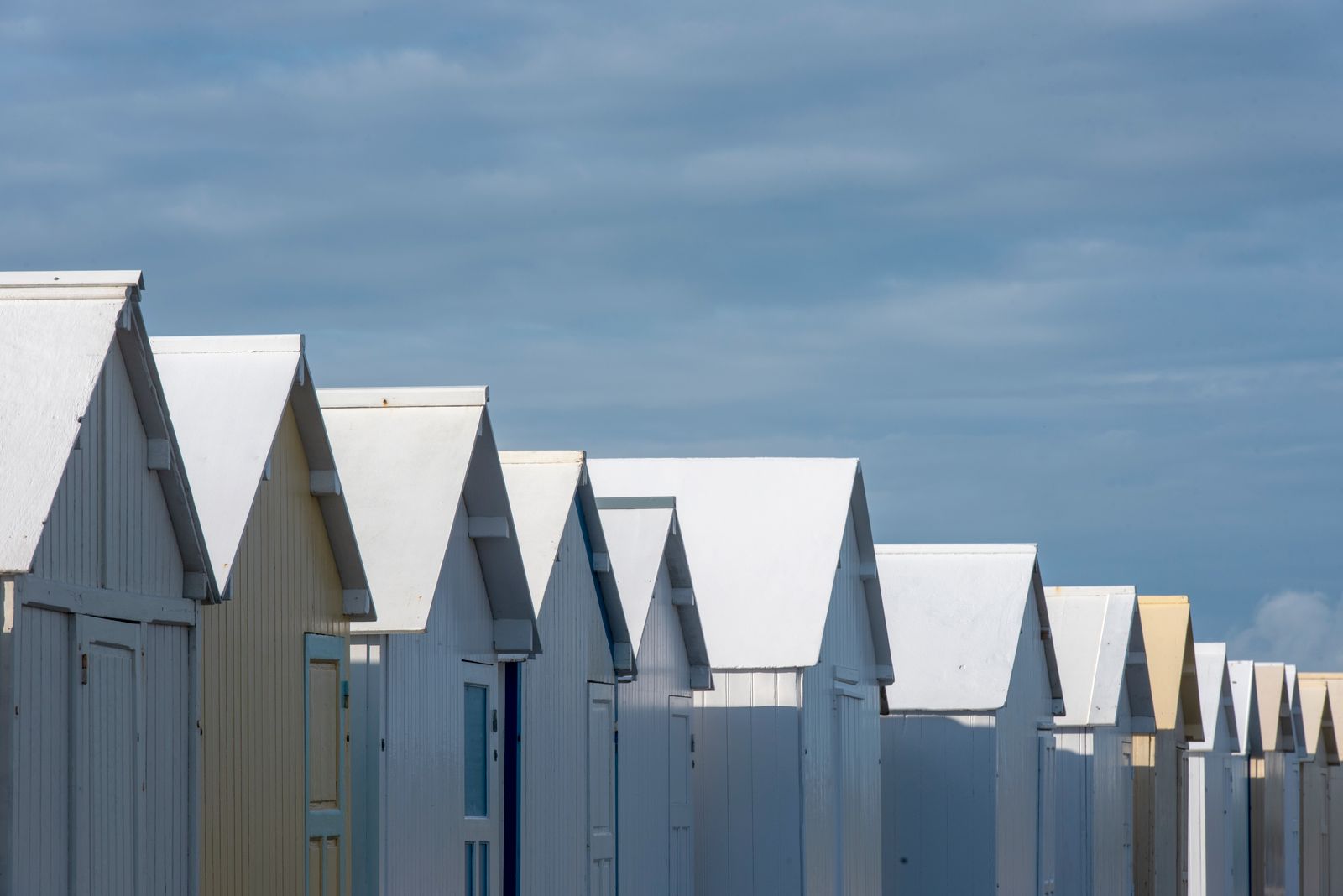 Une rangée de cabines de plage blanches et crème, alignées sous un ciel bleu pâle, chacune avec sa propre porte