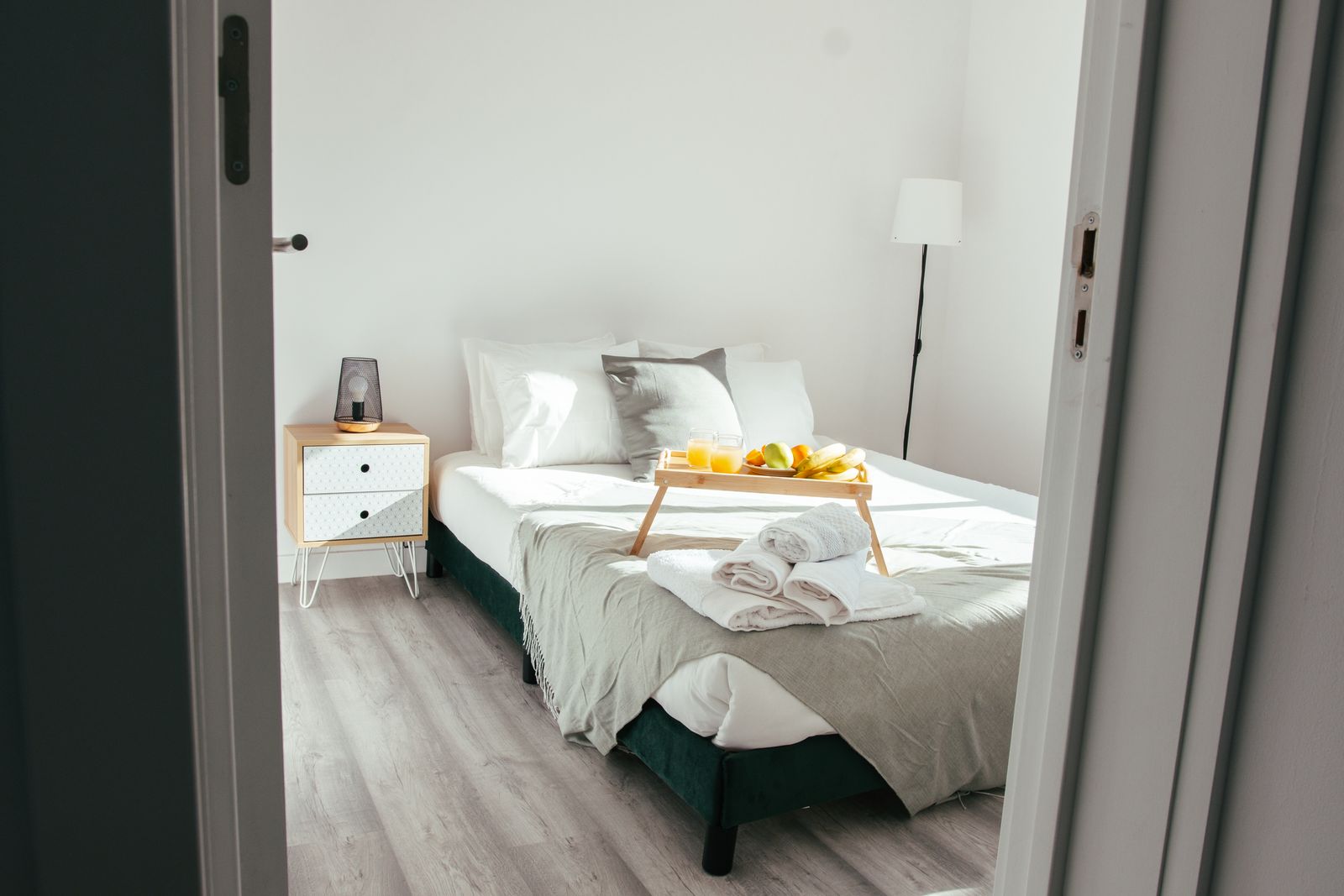 Une chambre blanche vue depuis la porte entrouverte, avec un plateau de fruits et des serviettes pliées posés sur le lit