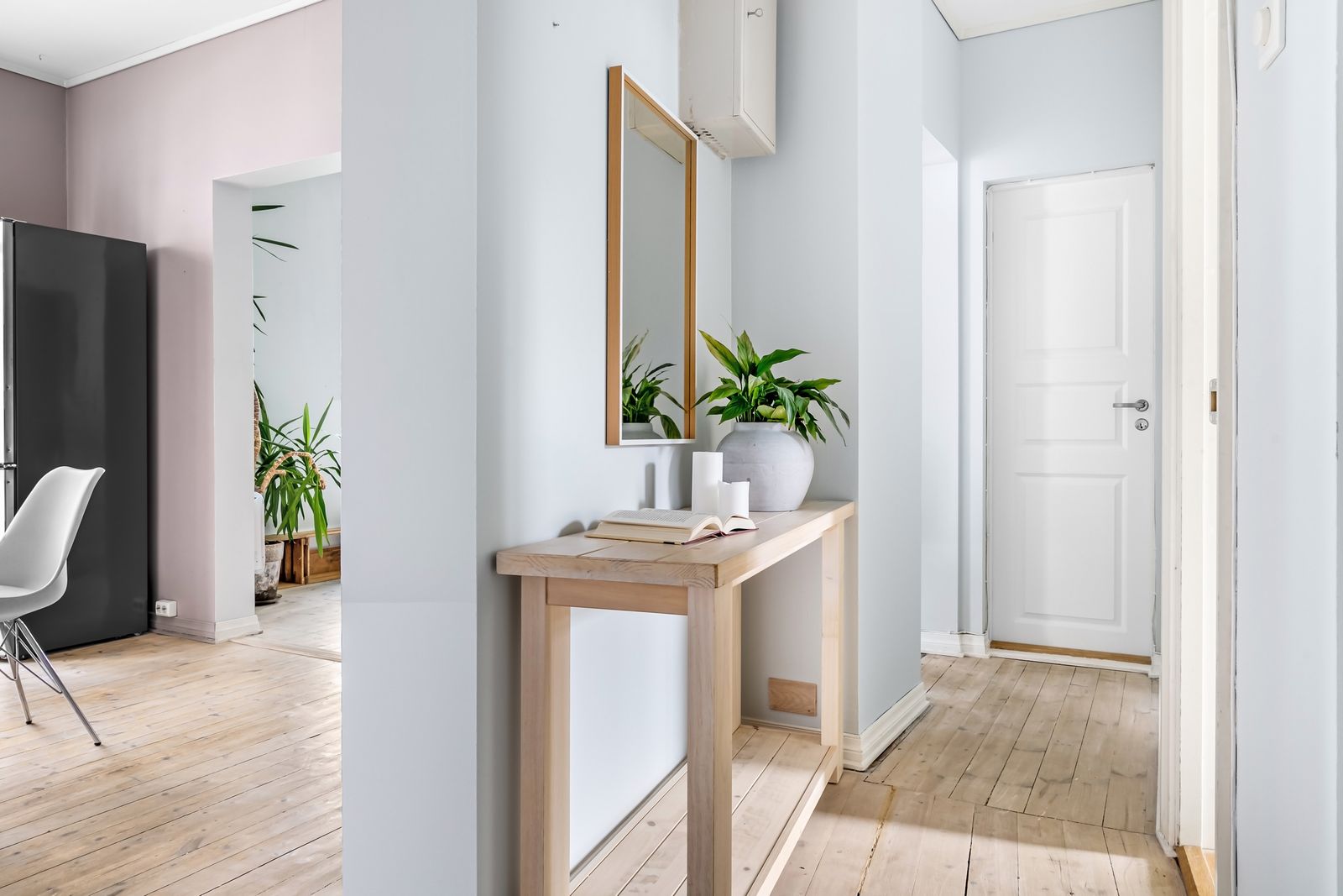 Une entrée claire au parquet blond, avec une console en bois sous un miroir, une plante verte, des bougies et un livre ouvert, la porte d'entrée blanche au fond