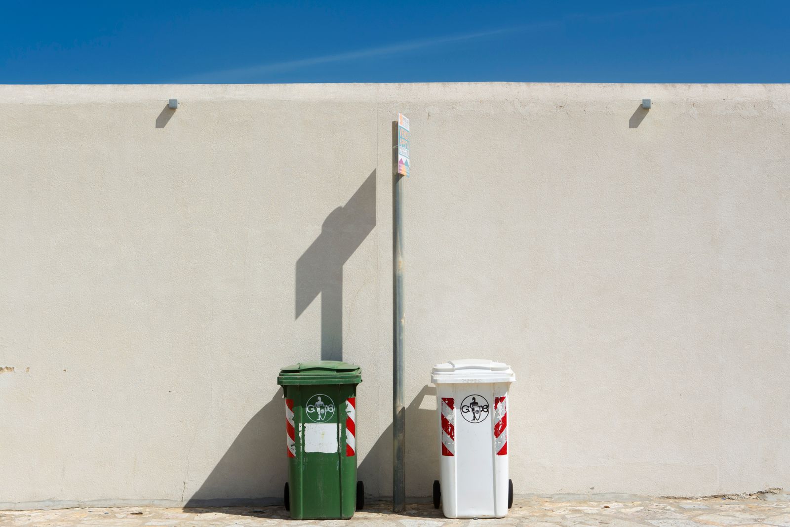 Deux bacs à roulettes, un vert et un blanc, posés contre un mur clair sous un ciel bleu