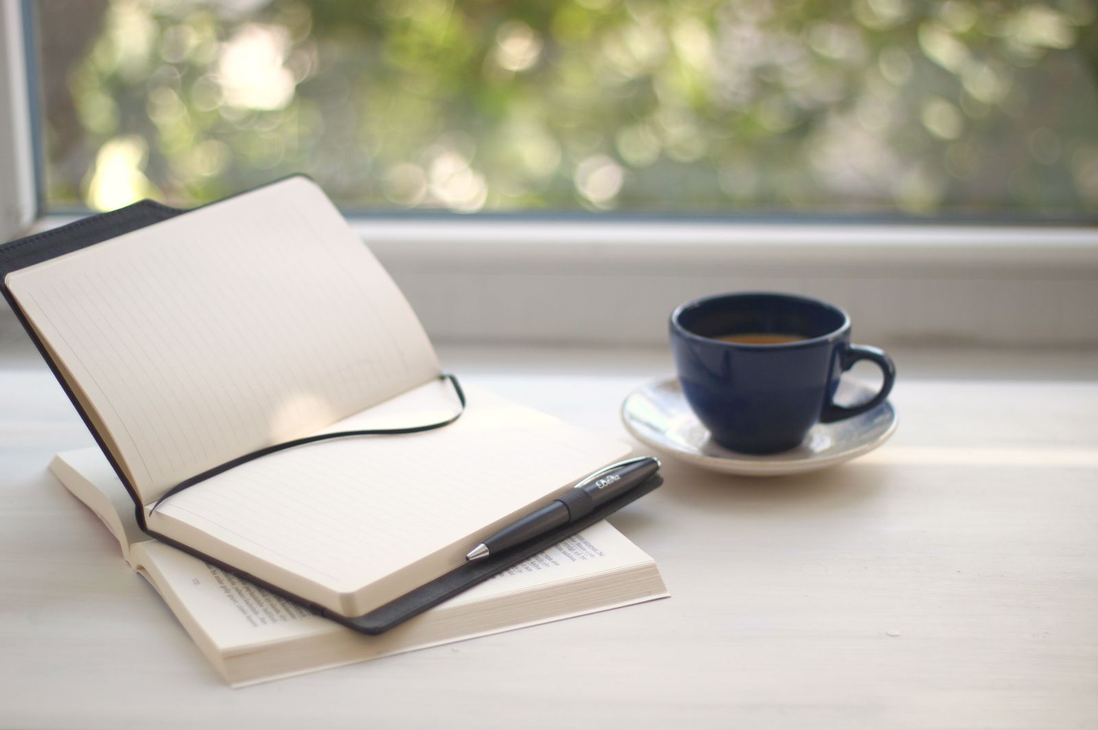 Un carnet ouvert aux pages blanches, un stylo et une tasse de café sur un rebord de fenêtre ensoleillé