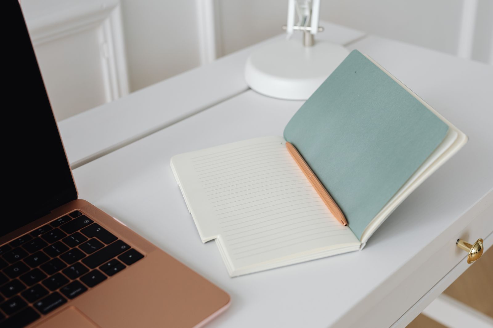 Un carnet ouvert et un crayon posés sur un bureau blanc, à côté d'un ordinateur portable et d'une lampe