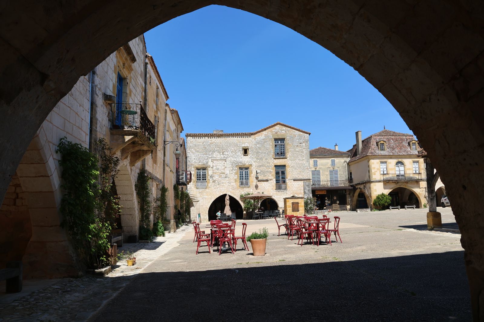 Des tables rouges de café sur la place ensoleillée d'un village de pierre, vues depuis une arcade