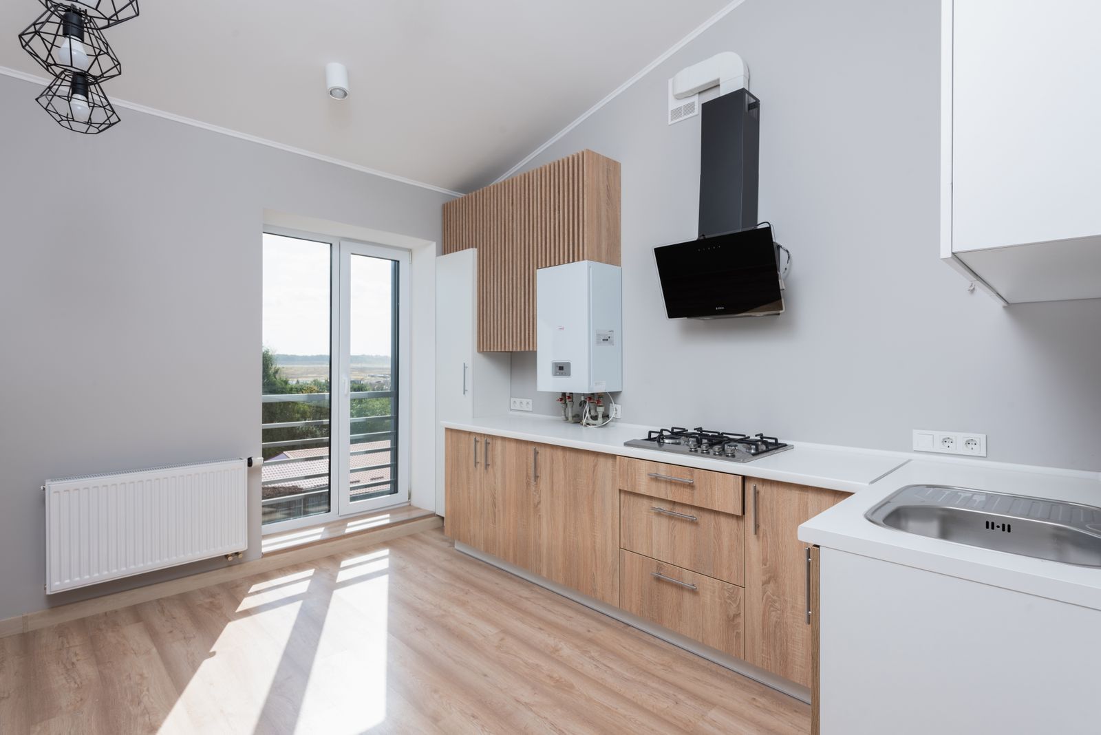 Une cuisine claire aux façades en bois, avec une plaque de cuisson, une hotte, une chaudière murale et une porte-fenêtre ouverte sur la campagne