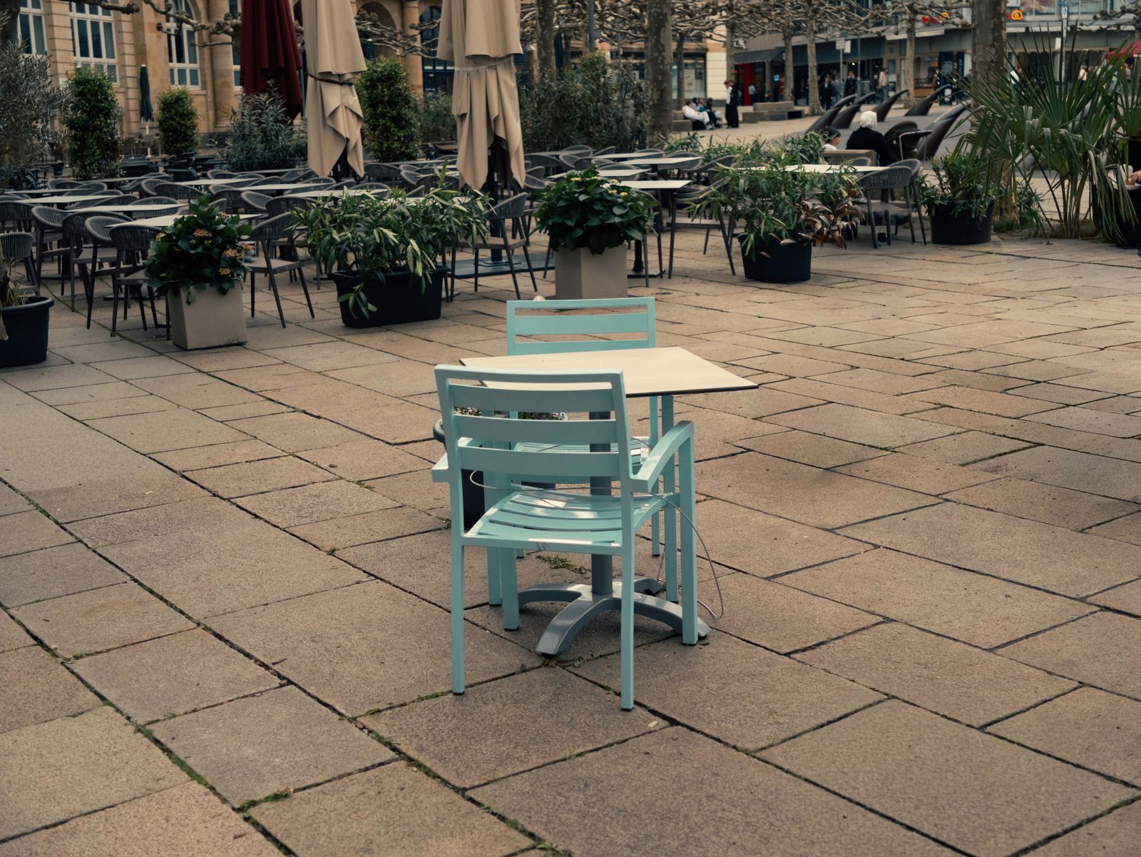 Une terrasse de café vide sur une place pavée, avec deux chaises bleu clair autour d'une petite table et des parasols fermés au fond