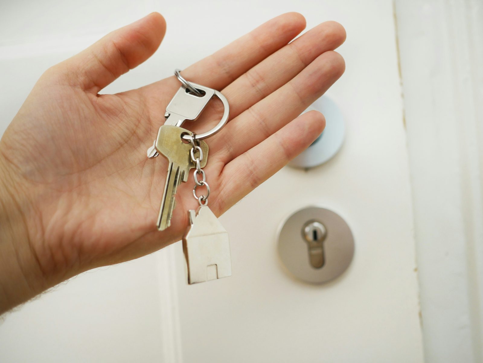 Une main ouverte tenant un trousseau avec un porte-clés en forme de maison, devant une porte blanche
