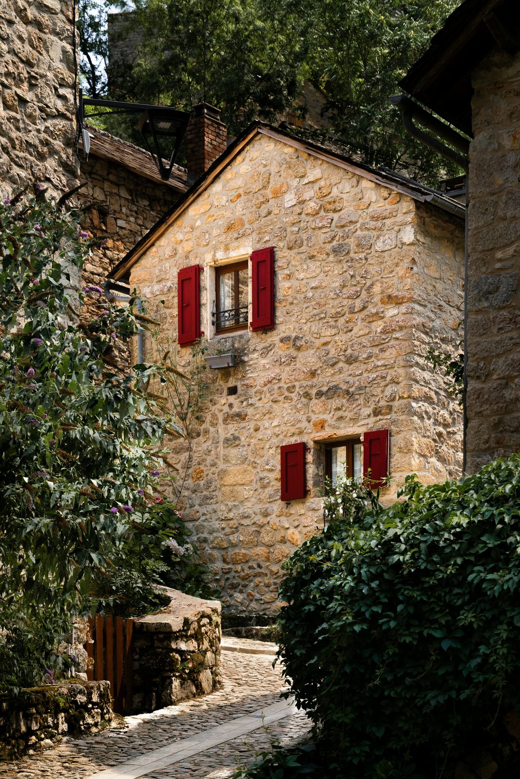 Une maison en pierre aux volets rouges, au bord d'une ruelle pavée bordée de verdure